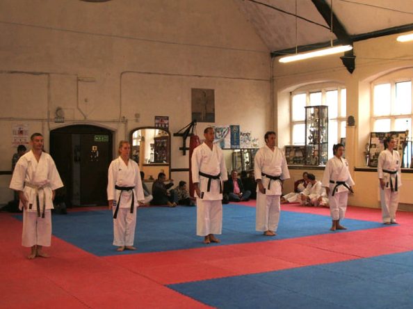 Black belts lined up on the red and blue mats in the club's church hall dojo, trophy cabinets behind